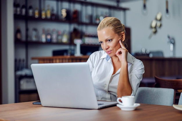 Une femme élégante blonde sérieuse est assise dans le café convivial et travaille sur un ordinateur portable