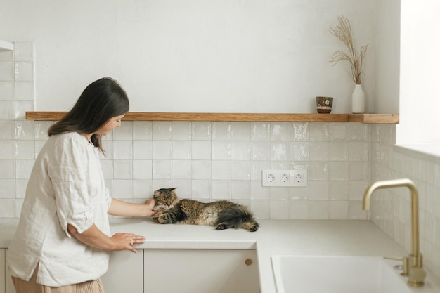 Femme élégante caressant son chat dans une nouvelle cuisine blanche minimale Femme au foyer nettoyant la cuisine après avoir emménagé avec un animal mignon dans une nouvelle maison moderne