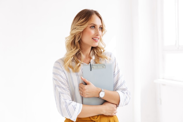 femme élégante caucasienne souriante et tenant le presse-papiers dans un bureau blanc