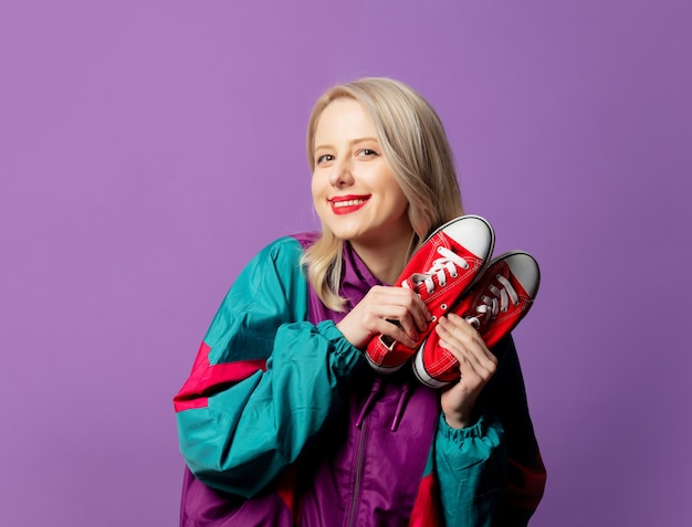 Femme élégante en coupe-vent des années 80 et lunettes de soleil rondes tient des chaussures rouges sur le mur violet