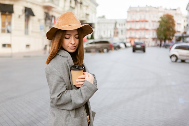 Une femme élégante du millénaire dans un manteau plus lisse et un chapeau de feutre branle une tasse de café lors de ses déplacements tout en marchant dans une ville européenne