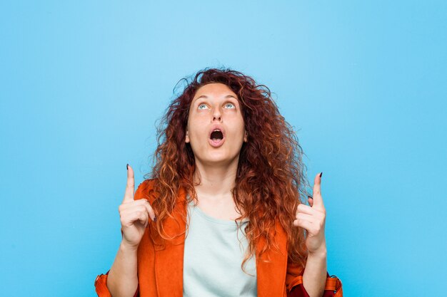 Femme élégante jeune rousse pointant vers le haut avec la bouche ouverte.