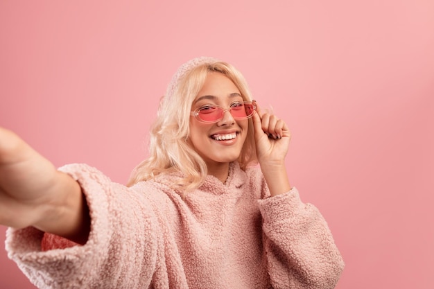 Femme élégante joyeuse faisant selfie portant des lunettes colorées et un sweat à capuche souriant à la caméra sur rose