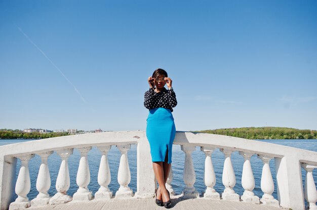 Femme élégante à lunettes, jupe bleue et chemisier noir posé en plein air sur le pont contre le lac