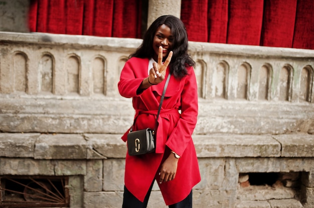 Photo femme élégante en manteau rouge posée contre de vieilles colonnes et montrer deux doigts signe