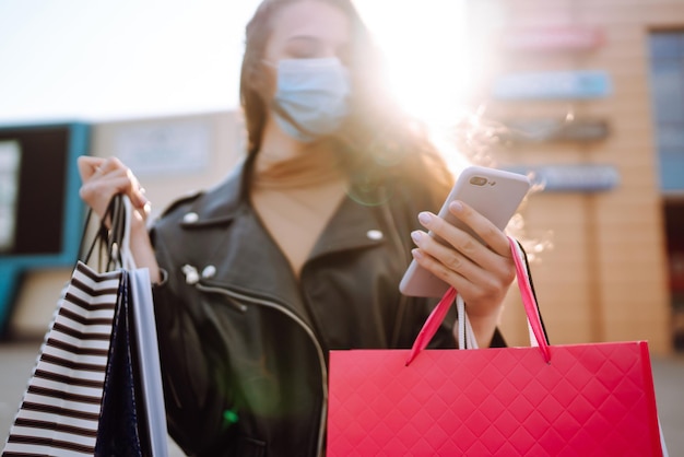Photo femme élégante portant un masque médical de protection avec des sacs à provisions à l'aide de son téléphone