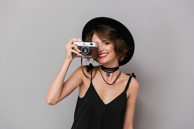 Femme élégante portant une robe noire et un chapeau photographier sur appareil photo rétro, isolé sur mur gris