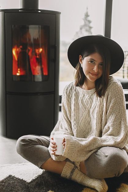 Photo femme élégante en pull et chapeau tricotés tenant une tasse de thé chaude et se relaxant devant une cheminée moderne