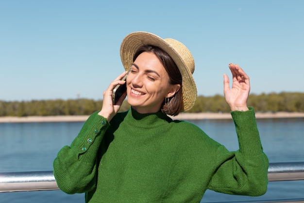 Femme élégante En Pull Décontracté Vert Et Chapeau En Plein Air Sur Le Pont Avec Vue Sur La Rivière à La Journée D'été Chaude Et Ensoleillée Parle Sur Téléphone Mobile