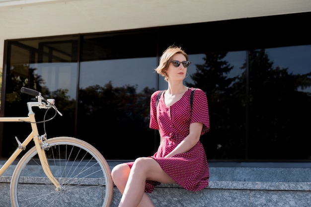 Femme élégante en robe à pois assis près d'un vélo rétro en zone urbaine. Belle jeune femme posant près d'un vélo vintage à l'extérieur