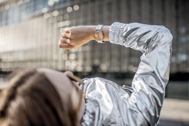 Femme élégante en veste argentée regardant l'horloge allongée à l'extérieur dans l'environnement moderne