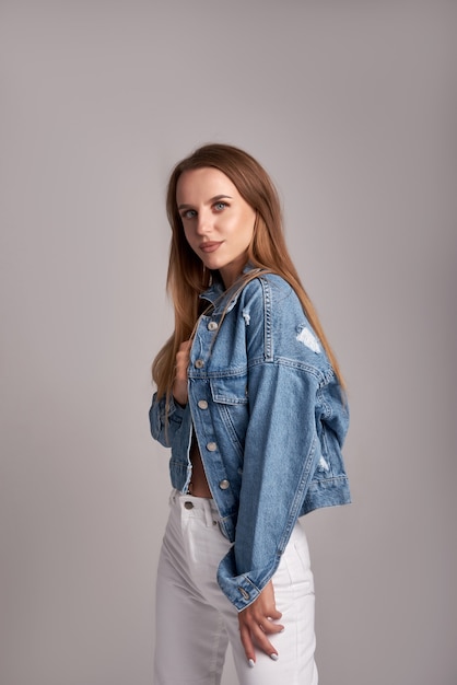 Femme élégante en veste en jean et jeans blancs posant sur fond gris