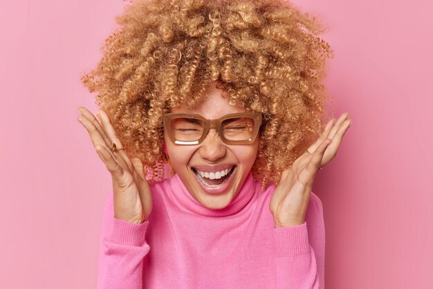 Une femme émotionnelle aux cheveux bouclés lève les mains près du visage se sent ravie s'exclame bruyamment se sent heureuse porte des lunettes transparentes et un col roulé isolé sur fond rose. Concept de réactions humaines