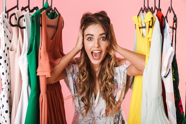 Femme émotionnelle en robe debout à l'intérieur d'une armoire pleine de vêtements isolés sur rose