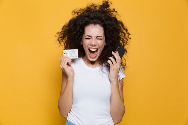 femme émotive des années 20 criant tout en tenant un téléphone portable et une carte de crédit isolée sur jaune