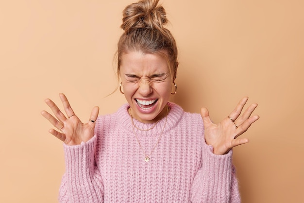 Une femme émotive aux yeux fermés réagit à quelque chose crie fort garde les paumes levées a les cheveux peignés en chignon porte un pull tricoté entend des gestes de nouvelles incroyables se tient activement sur fond beige