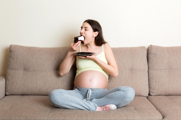 Une femme enceinte affamée mange un morceau de gâteau savoureux en se relaxant sur le canapé à la maison Envies sucrées pendant la grossesse