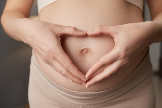 Une femme enceinte aimante et inconnue avec le ventre nu se tient à l'intérieur de la maison en forme de cœur avec les mains sur son ventre exprimant la douceur en portant des vêtements beige