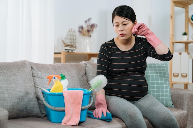 Une femme enceinte asiatique inconfortable éponge la sueur avec la main tout en se reposant sur un canapé dans le salon après avoir fait des travaux ménagers lors d'une journée d'été surchauffée.