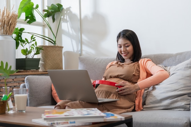 Une femme enceinte asiatique sourit et assise sur le canapé, écoutant de la musique et utilisant un ordinateur portable avec un sentiment de bonheur et de détente.