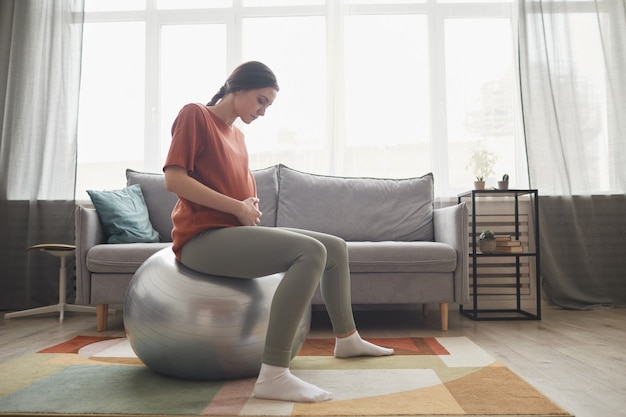 Femme enceinte assise sur un ballon de fitness et faisant de l'exercice pendant l'entraînement sportif à la maison