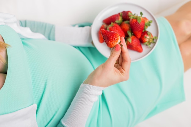 Femme enceinte assise sur un canapé et mangeant des fraises dans un bol