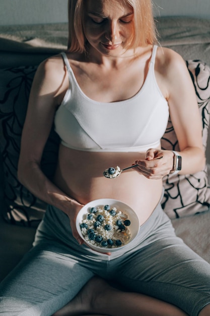 Femme enceinte assise sur le canapé et tenant une assiette avec de la ricotta et des myrtilles