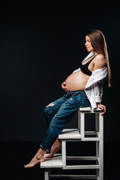 Femme enceinte assise sur une chaise échelle dans un studio sur fond noir.