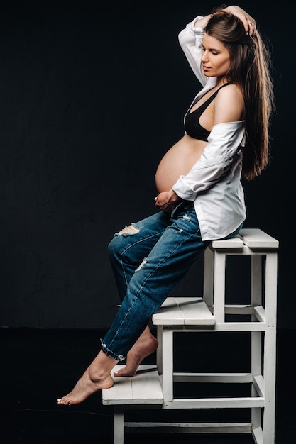 Femme enceinte assise sur une chaise échelle dans un studio sur fond noir.