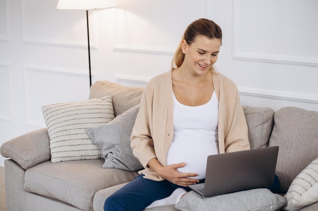 Femme enceinte assise sur l'entraîneur à l'aide d'un ordinateur portable et tenant des écouteurs par le ventre