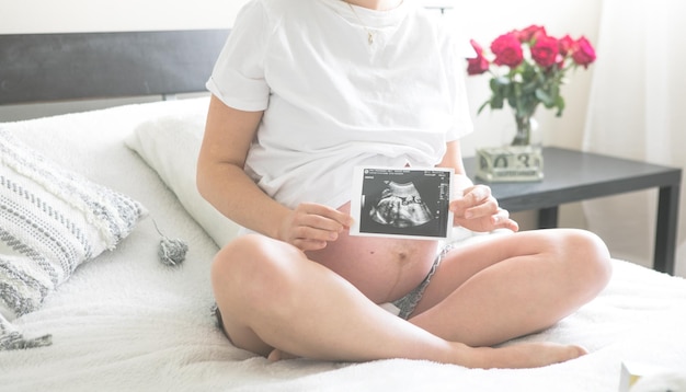 Femme enceinte assise sur un lit tenant une image radiographique de son bébéLe concept de grossesse, de maternité et de soins prénataux Maman avec une nouvelle vie