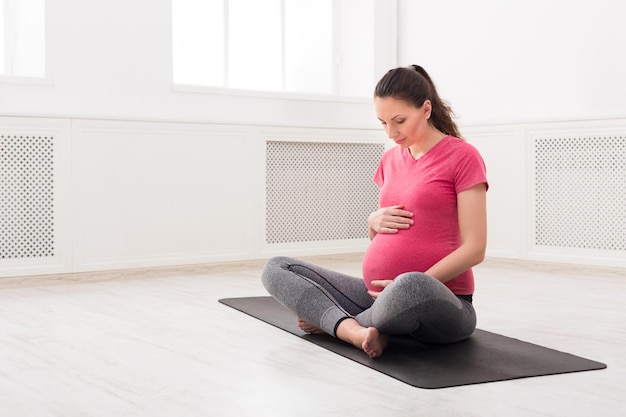 Femme enceinte assise en posture de lotus, caresse son ventre. Une femme enceinte se repose tout en pratiquant le yoga à la maison, la méditation, la relaxation, le concept de grossesse saine, l'espace de copie