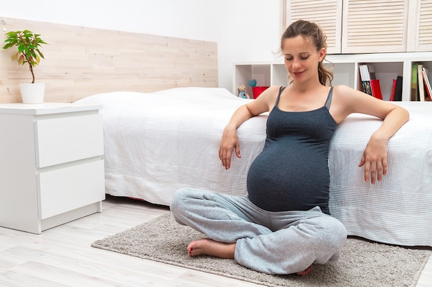 Femme enceinte assise sur le tapis avec les jambes croisées et se penchant en arrière sur le lit à l'intérieur