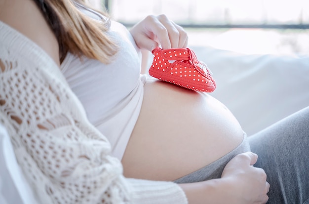Femme enceinte assise et tenant des chaussures de bébé rouge mis sur son ventre à la maison
