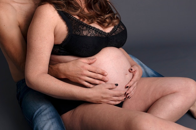 Une femme enceinte assise tient la main de son mari dans le gros ventre. Couple attend un bébé dans un fond gris.