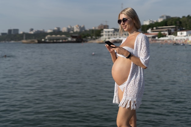 Femme enceinte au bord de l'océan. Tapez un message sur le téléphone. Concept de vacances pendant la grossesse