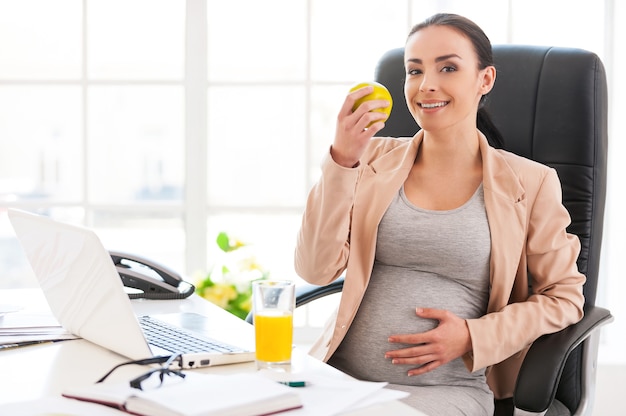 Femme enceinte au bureau. Belle femme d'affaires enceinte mangeant une pomme et souriant à la caméra alors qu'elle était assise sur son lieu de travail