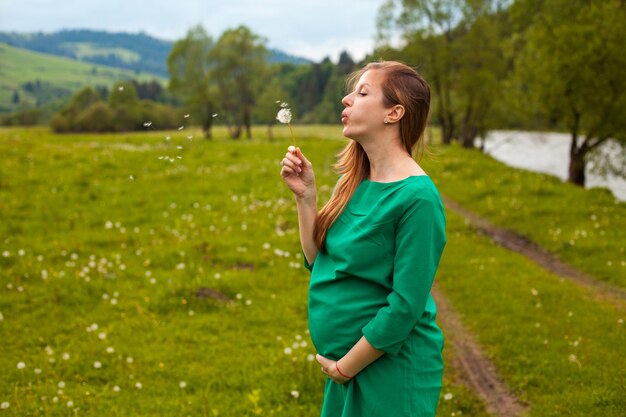 Photo une femme enceinte aux cheveux bruns dans une belle tunique verte souffle un air de pissenlit sur le fond de la nature avec des arbres verts