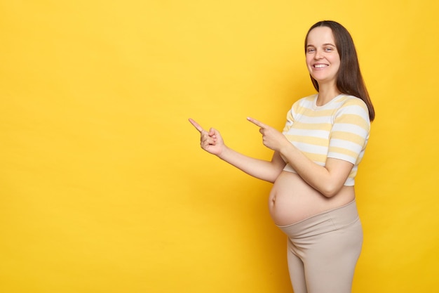 Une femme enceinte blanche souriante et joyeuse en vêtements décontractés posant isolée sur un fond jaune pointant vers l'espace de copie pour la publicité ou le texte promotionnel