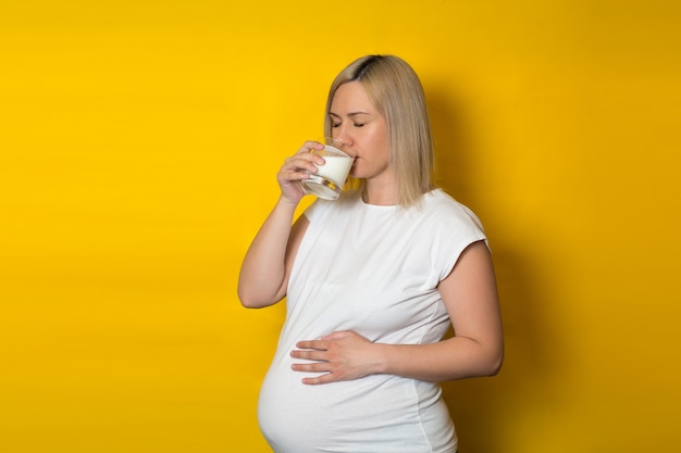 Femme enceinte, blonde buvant du lait dans un verre, tenant sa main sur son ventre, sur un mur jaune