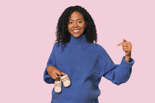 Une femme enceinte avec des chaussures de bébé dans un studio, une personne montrant de la main un espace de copie de chemise, fière.