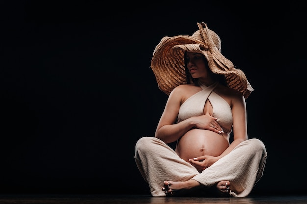 Une femme enceinte dans un chapeau de paille est assise sur le sol dans des vêtements beiges dans un studio sur fond noir.