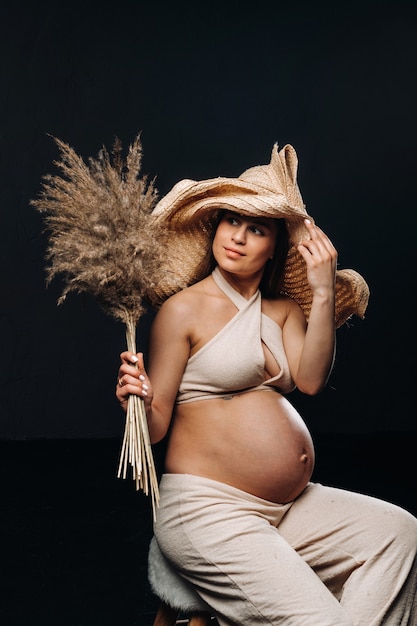 Femme enceinte dans un chapeau de paille avec des vêtements beiges avec un bouquet dans ses mains en studio sur fond noir