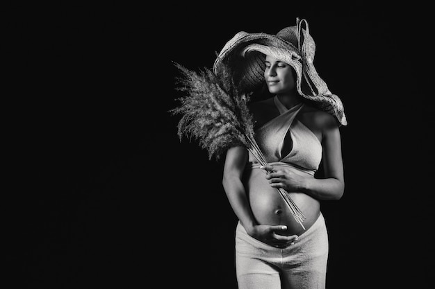 Femme enceinte dans un chapeau de paille avec des vêtements beiges avec un bouquet dans ses mains en studio sur fond noir