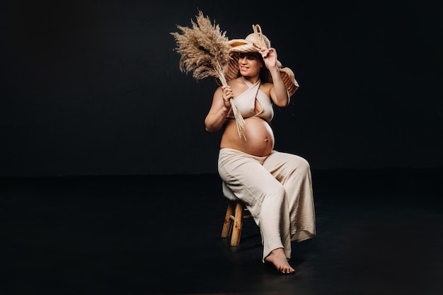 Femme enceinte dans un chapeau de paille avec des vêtements beiges avec un bouquet dans ses mains en studio sur fond noir.