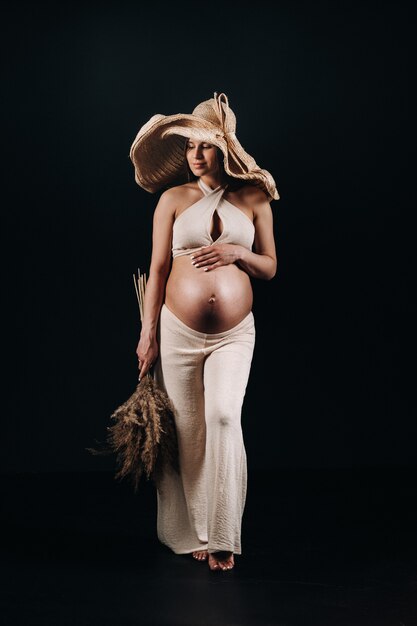 Femme enceinte dans un chapeau de paille avec des vêtements beiges avec un bouquet dans ses mains en studio sur fond noir.