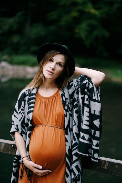 Photo femme enceinte dans un chapeau se penche sur la balustrade d'une jetée sur le portrait du lac