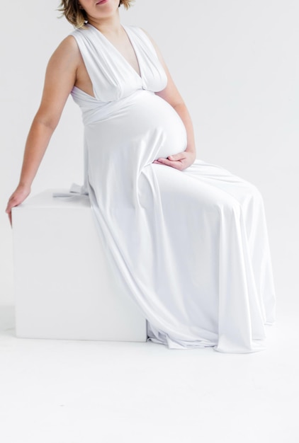Une femme enceinte dans une longue robe de soie blanche étreint son ventre et pose sur un fond blanc assis sur un cube blanc