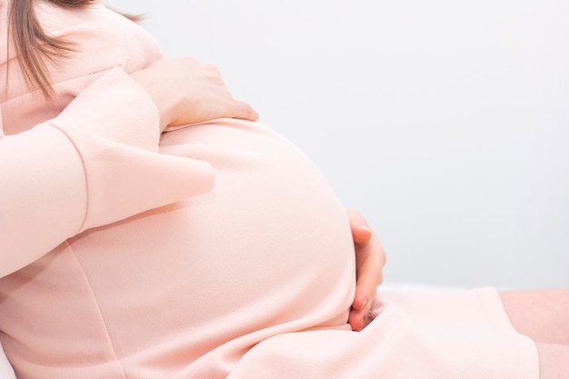 Une femme enceinte dans une robe tient ses mains sur son ventre sur un fond clair. Belle grossesse photo humeur tendre.