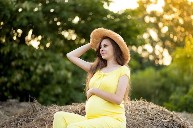 Une femme enceinte dans des vêtements sombres et un chapeau se trouve dans un champ sur de la paille en été, une promenade de fille enceinte dans la nature